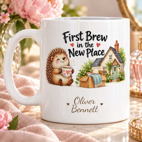 First Brew in the New Place – Personalised Mug — cheerful cartoon hedgehog holding a steaming teacup beside a tiny new cottage and unpacked box